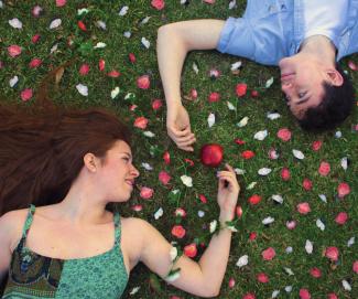 Actors from 'The Diary of Adam and Eve' lay on grass peering at each other. Flower pedals cover the ground and an apple is between their hands.
