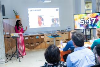 A young woman presents a business pitch in front of two monitors while a seating group of people watch and listen