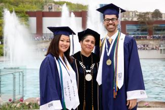 Shirley M. Collado with two students in caps and gowns in front of fountains