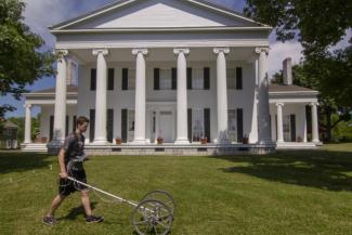 A man pushes a rolling machine in front of a white, Greek Revival style mansion