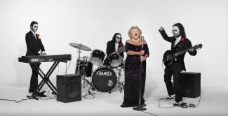An older woman in black sings while men in black suits and skull makeup play instruments.