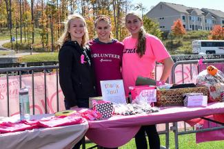 Three young women stand together and smile while standing outdoors at a table covered in raffle prizes.