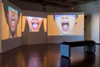 Three blindfolded faces with mouths wide open are projected on individual screens on gallery wall. 