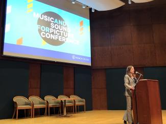 Woman speaking at podium with a screen behind her that says Music and Sound for Picture Conference