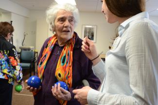 An elderly woman is holding two small blue balls standing up and speaking with a student in a white shirt