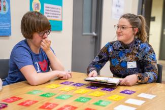 A student in a colorful shirt and glasses is sitting at a table with a young client. There are multi-colored cards spread out on the table in what looks like a game. The two are having a conversation