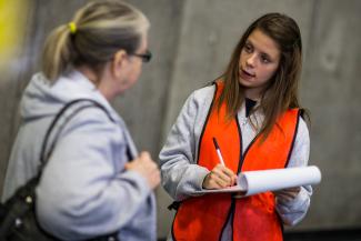 A student in an orange safety vest holding a clipboard is speaking to a person asking questions and writing them down on the clipboard.