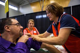 A person in a purple shirt is seated and pulling up the sleeve so that a person with a flu shot is able to administer the shot in the arm. A student stands in the background making notes.