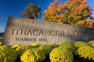 A stone marker with the words 'ithaca college' on them, surrounded by bushes, shrubbery, and autumn trees in the background