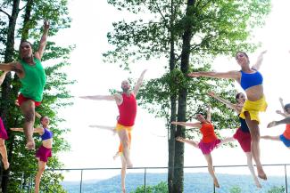 An outdoor shot of dancers in brightly-colored costumes leaping in front of a scenic mountain background.