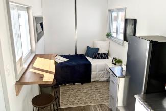 Inside of a shipping container, showing a bed, desk, and fridge