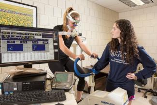 One student is on an exercise bike with a mask attached to her face breathing into it while exercising while the other student in a blue shirt is standing next to them observing vital signs with a computer screen.