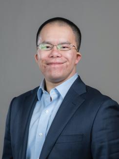 Photograph of Jerome Fung in a navy blue jacket standing in front of a gray background.