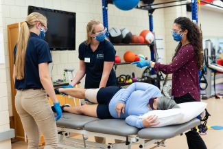 Image shows a patient lying on a their side on a plinth with an ace bandage around their left knee. One student therapist carefully stretches the injured knee while a second student watches closely. A supervisor gestures instructions. All four people are wearing masks. There is a shelf with weights and exervise balls in the background.