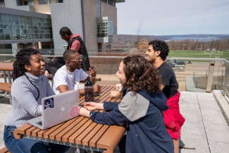 four students sitting at a table on a deck outside engaging in conversation