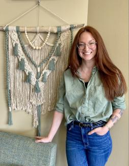Danni is smiling and stands with a macrame wall hanging she designed. She has glasses, long red hair, and a green button down shirt on.
