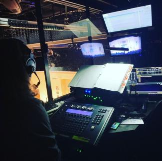 A Stage Management student calling a show in the booth. Their script is open and small monitors show the production on stage.
