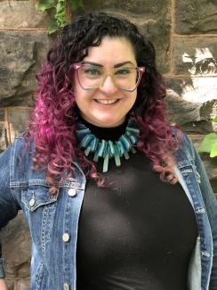 A white woman with purple hair and rainbow glasses stands smiling in front of a stone wall