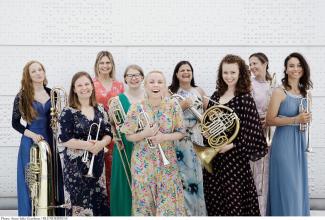A group of women wearing colorful dresses, standing, laughing, and holding brass instruments