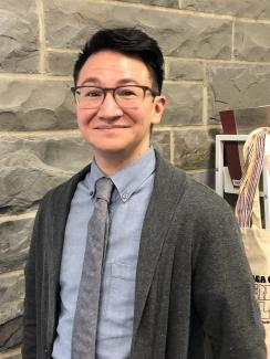 Lee is standing and smiling in front of a stone wall. They are a masculine-presenting, light-skinned individual with short brown hair and wearing dark-rimmed glasses, a blue button-down shirt, a grey cardigan, and a grey tie.
