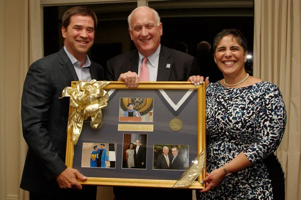 Two men and a woman pose as they hold a framed piece that showcases several photos and a medal. 