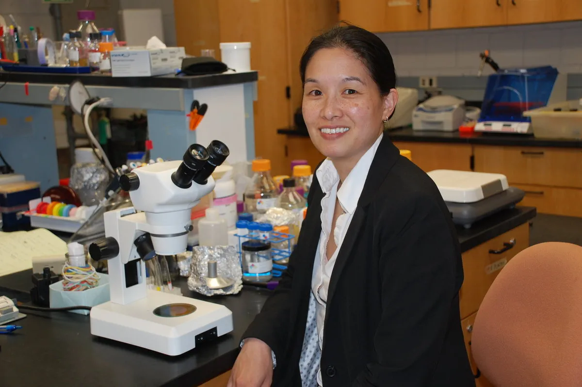 A woman sits and poses at a lab table, surrounded by scientific equipment. 