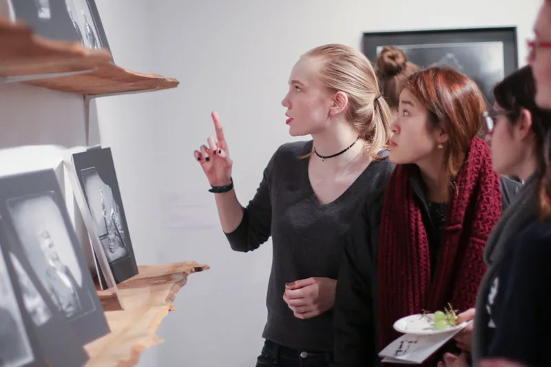 A group of female students peer at artwork propped up on wooden shelves. 