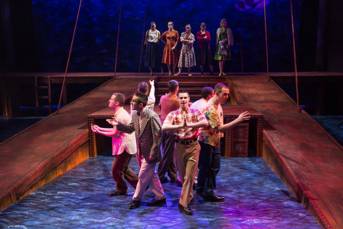 A group of men sing and dance on stage while women pose behind them