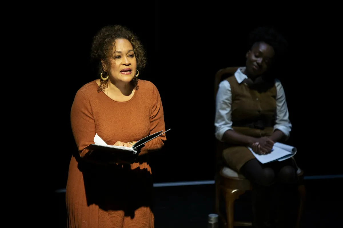 A woman in a spotlight reads aloud while another watches