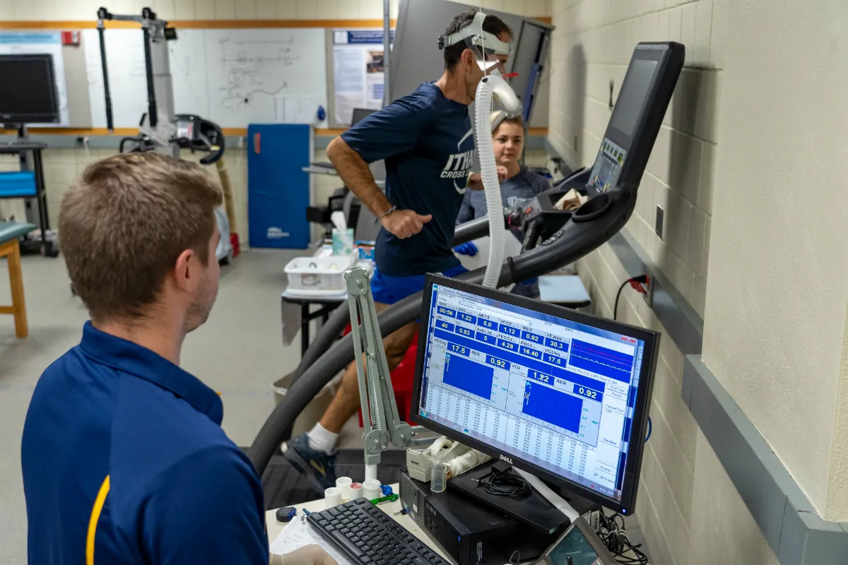 A man runs on a treadmill with a mask on as two others watch