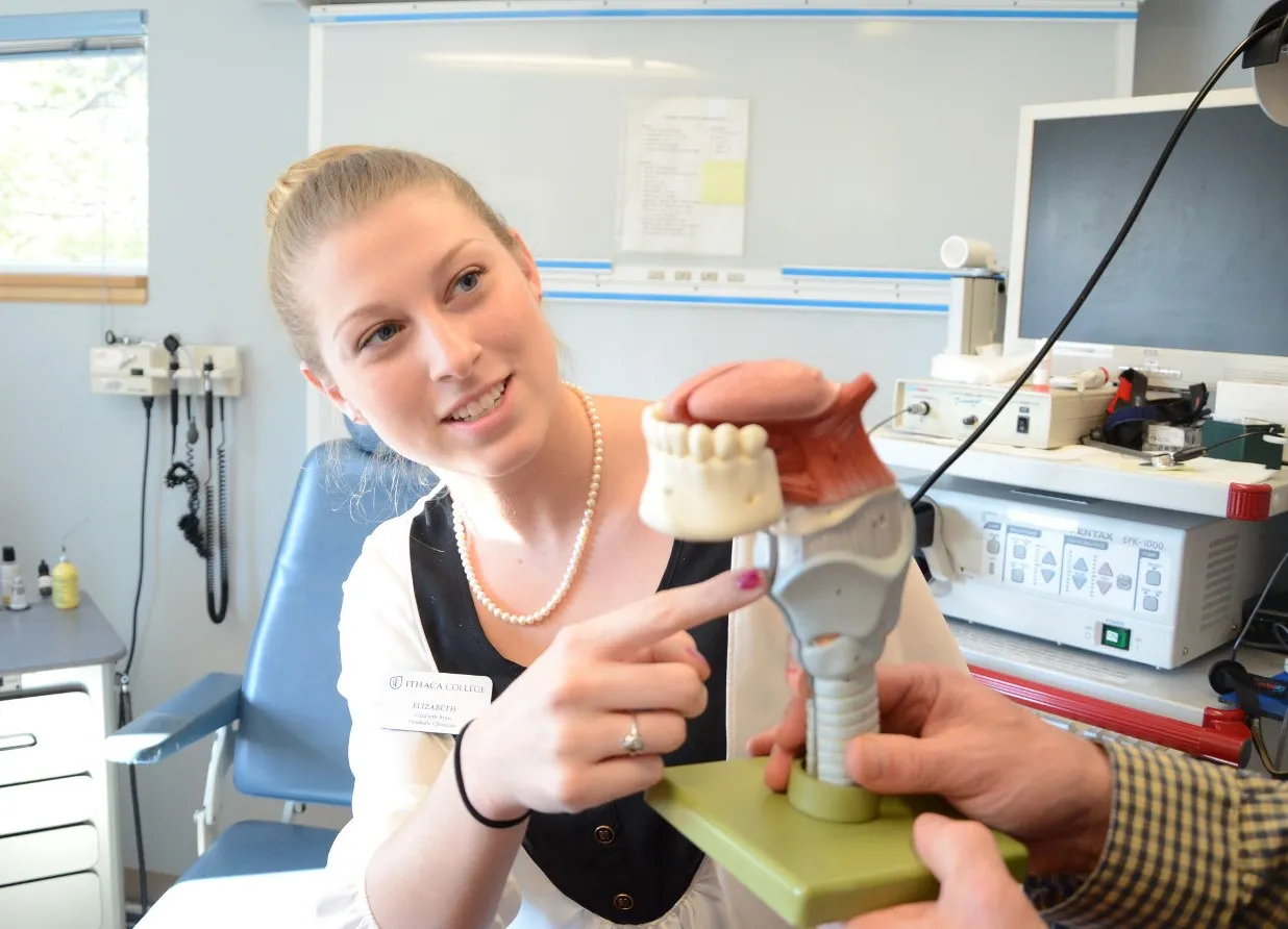 A student in a white shirt wearing pearls in seated in an examining chair pointing at an anatomical model of the throat.