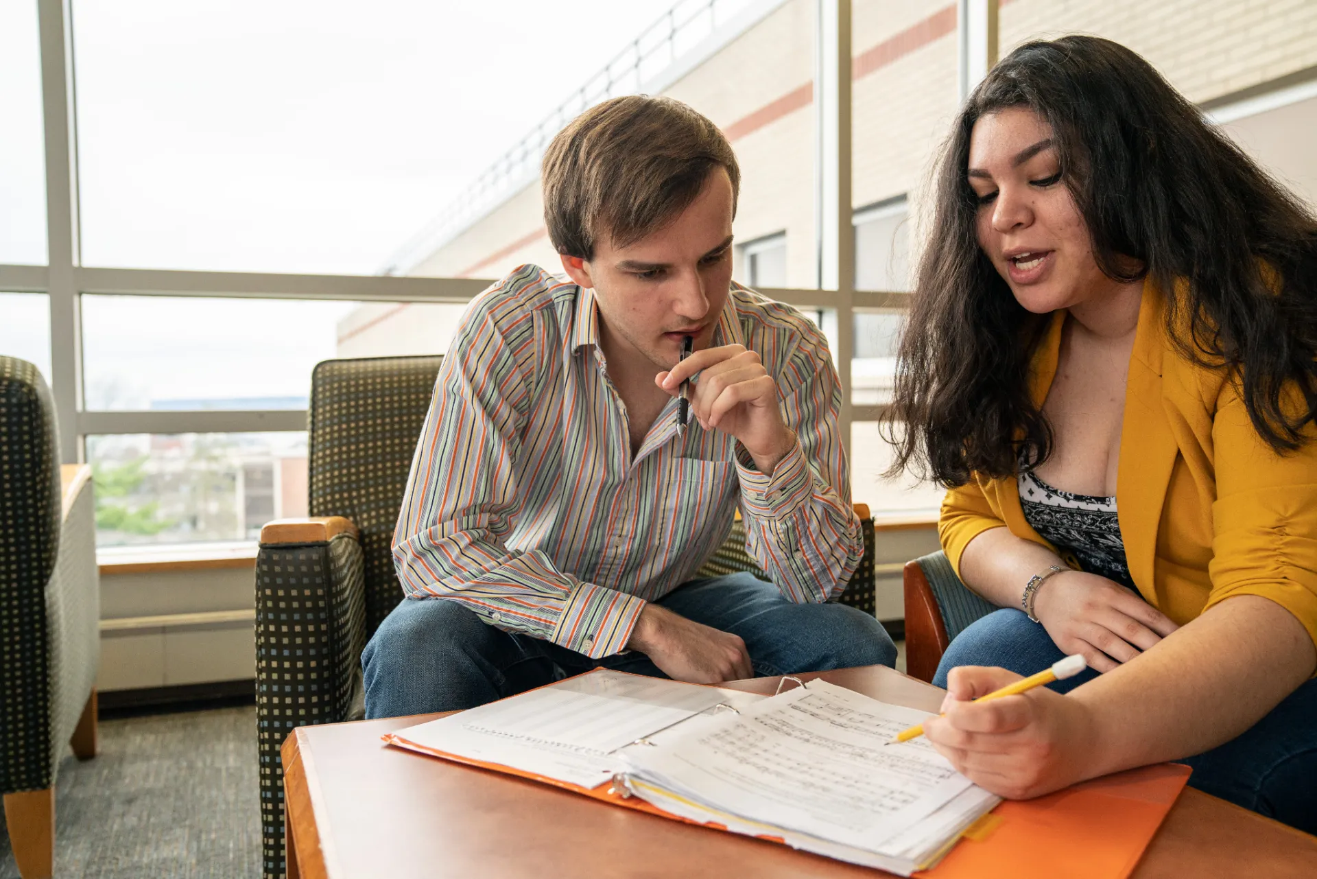 Two students sitting together, both looking at some papers on a table. One student points to the paper, appearing to explain something to the other, who looks deep in thought..