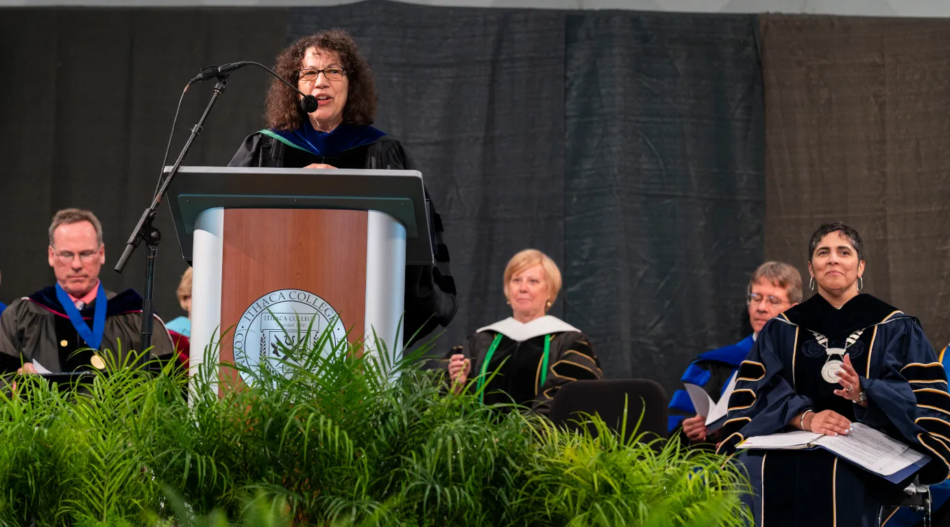 This is a photo of Linda Petrosino, Dean standing at a podium with other faculty members seated behind her. She is congratulating the new graduates.