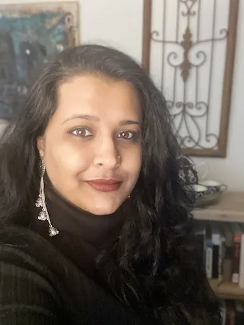 Headshot of south asian woman, center left, with wall hangings and bookshelves in the background