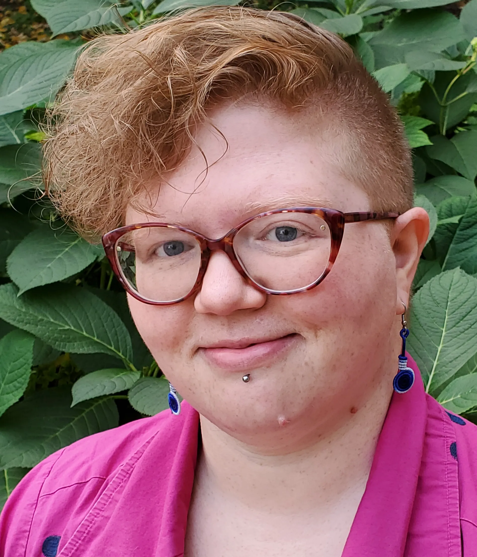 Person with a buzzed haircut wearing glasses, a pink dress, and a labret piercing, smiling at the camera.