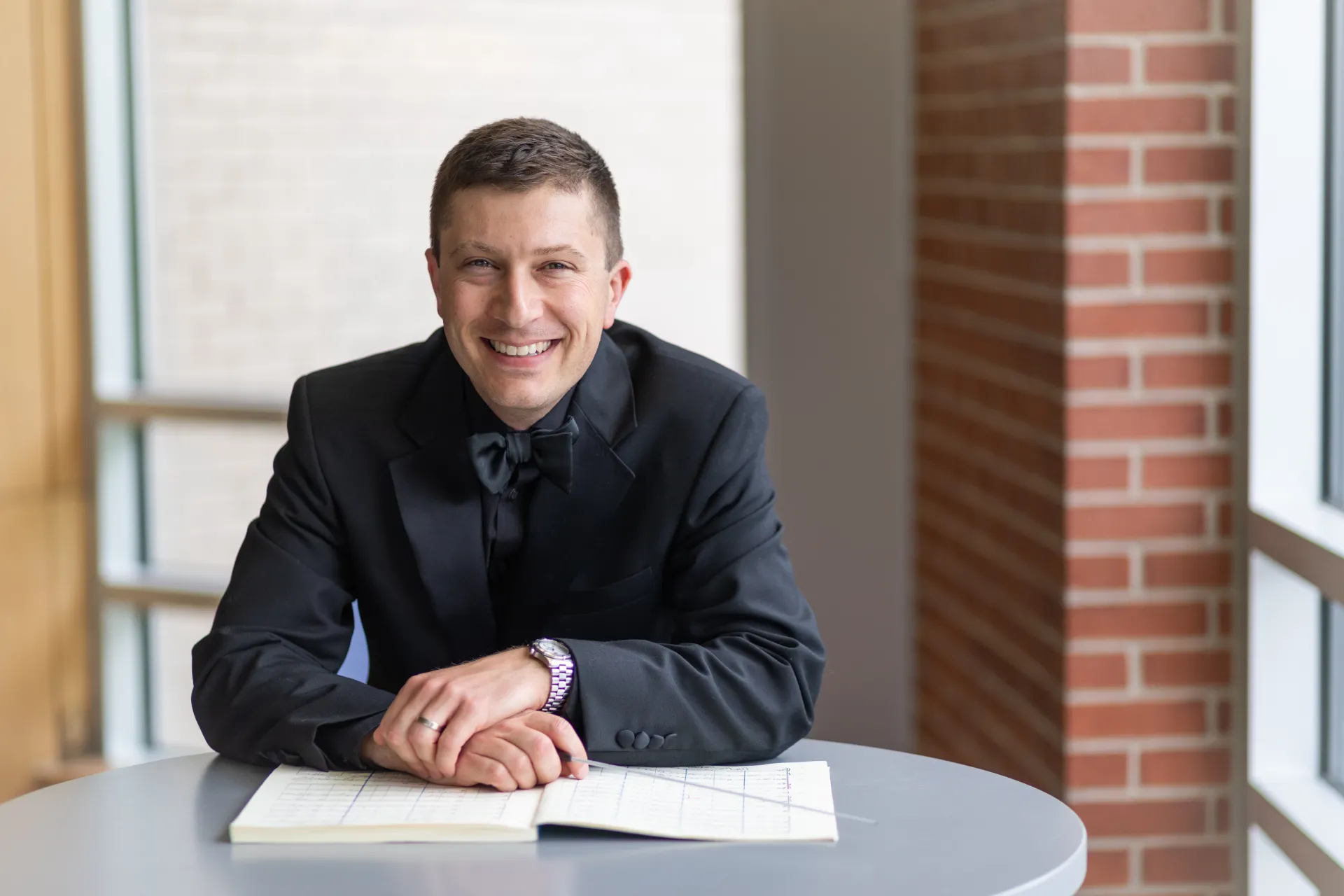 Headshot of Aaron Burgess, seated with score and baton, dressed in concert black, smiling from ear to ear!