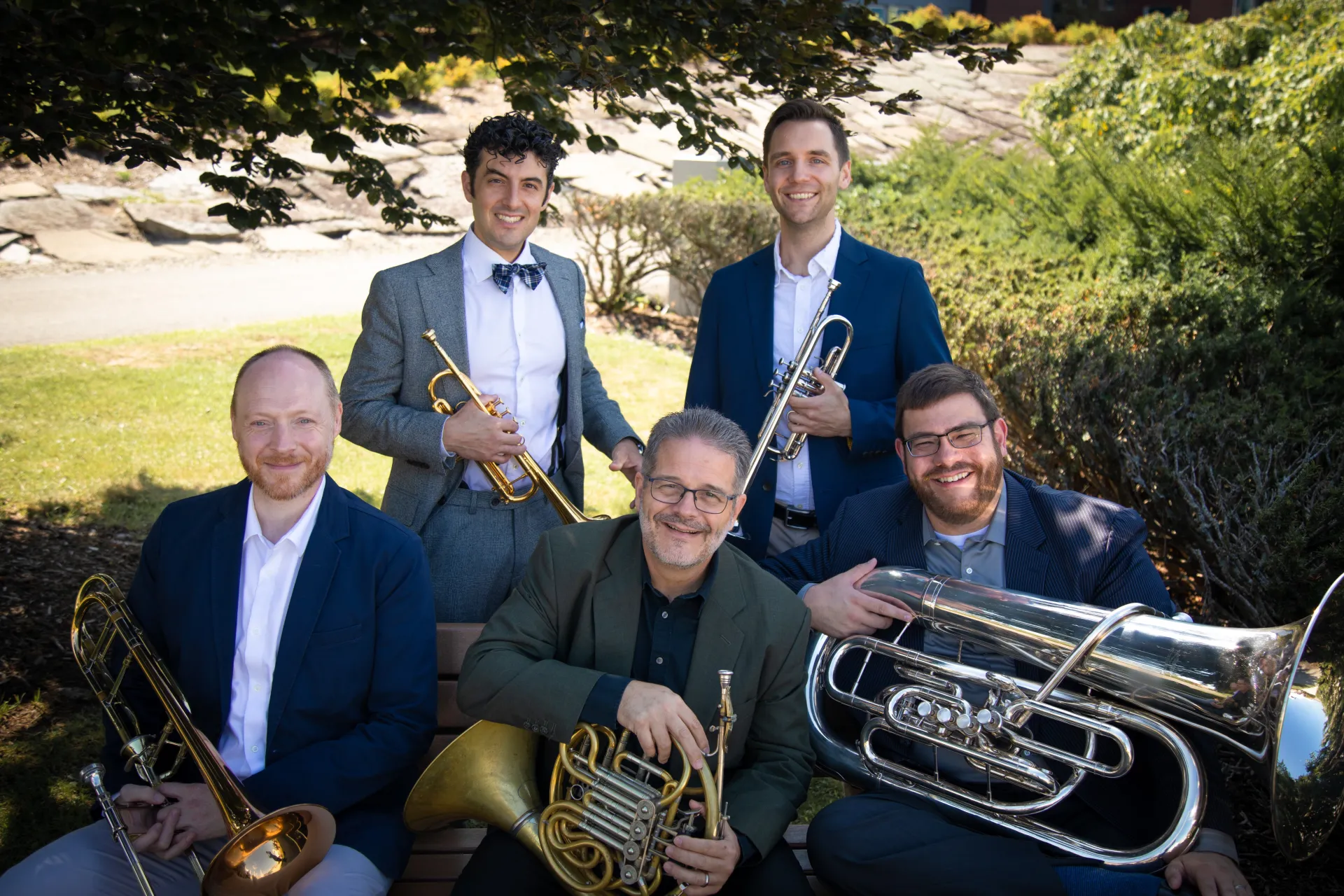 Five musicians holding instruments sitting outside under a tree