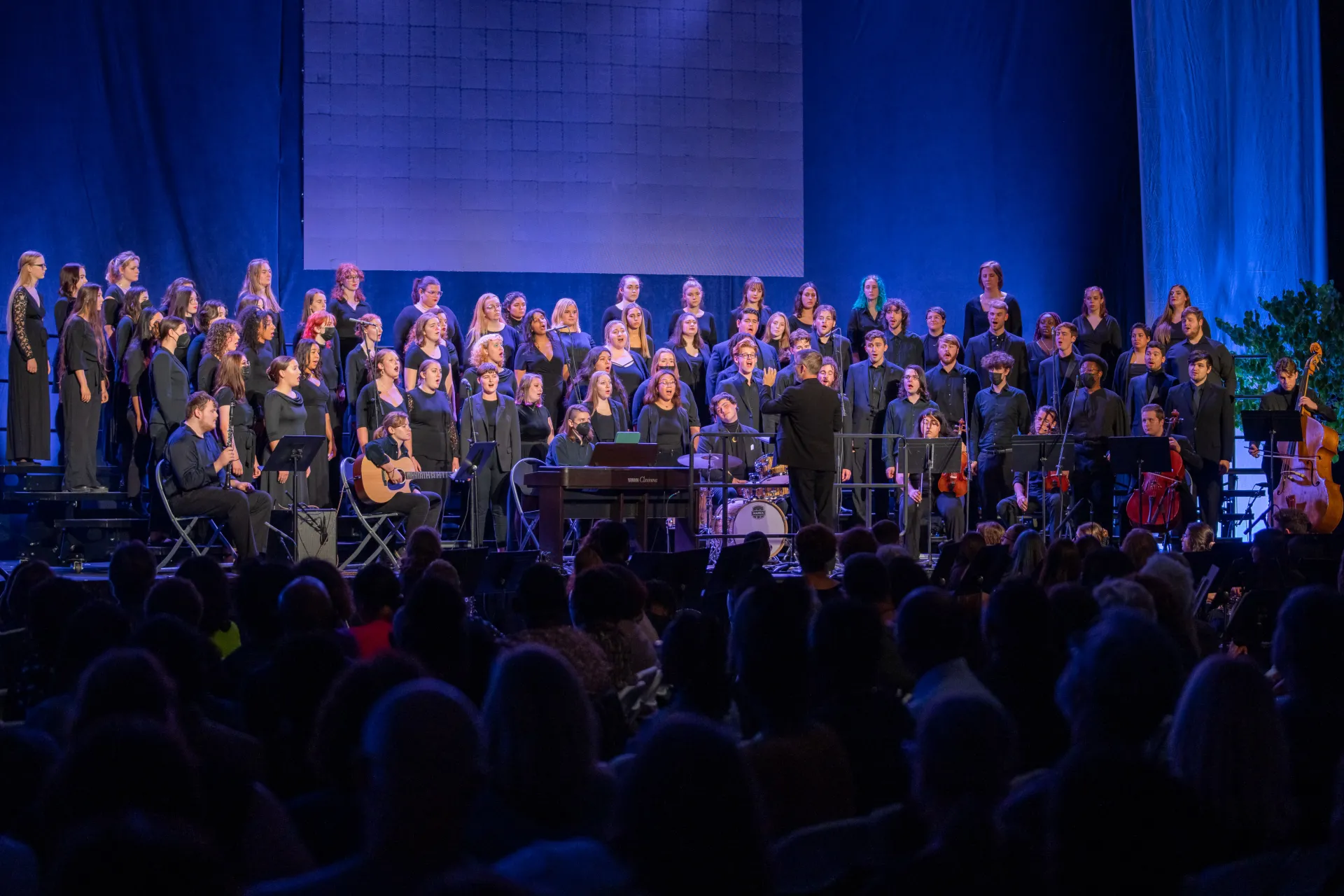 A pianist plays the piano with a chorus group singing at a concert.
