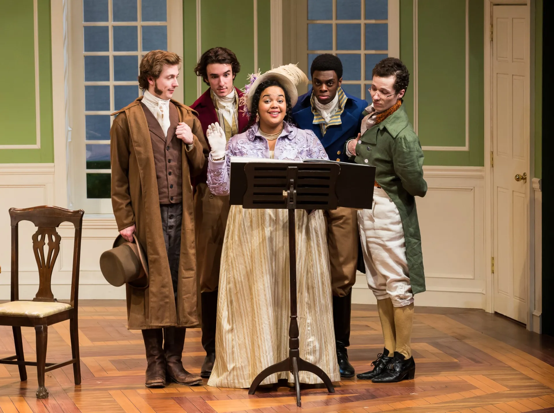 Five actors in 1800s costumes stand around a music stand. 