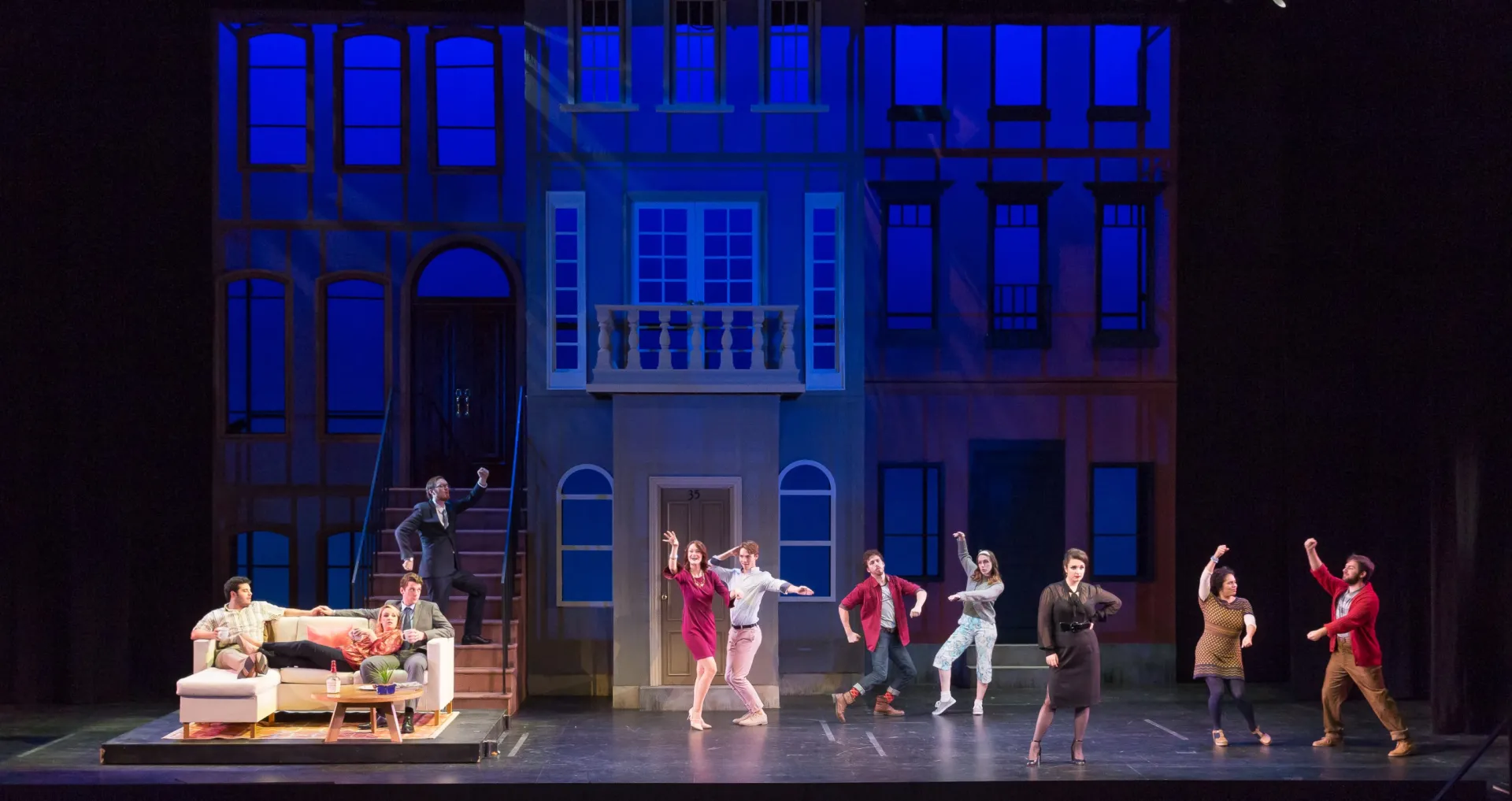 Three actors sit on a couch on one side of the stage, while three pairs of actors dance on the right. The set is a New York City apartment facade.