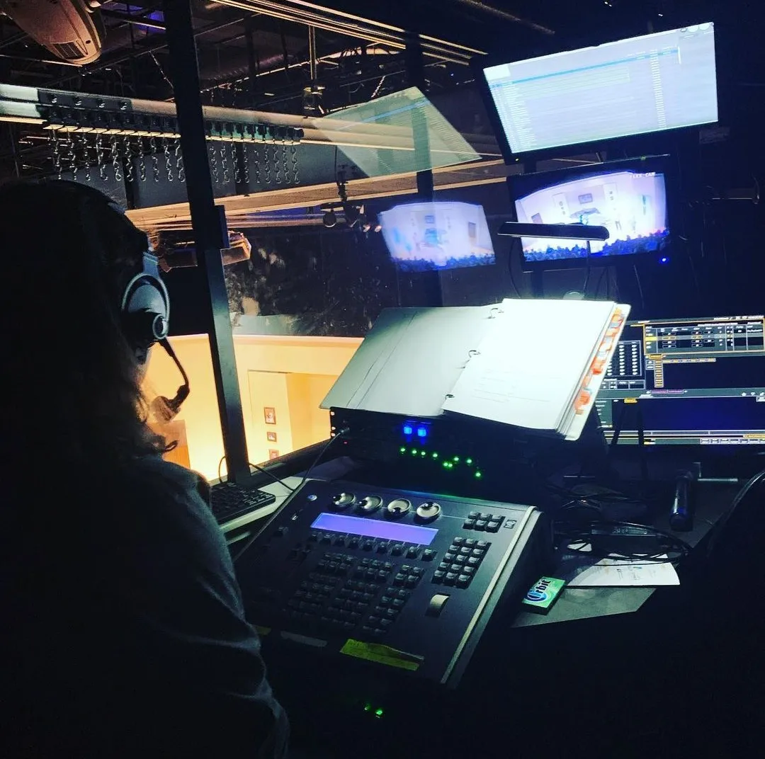 A Stage Management student calling a show in the booth. Their script is open and small monitors show the production on stage.