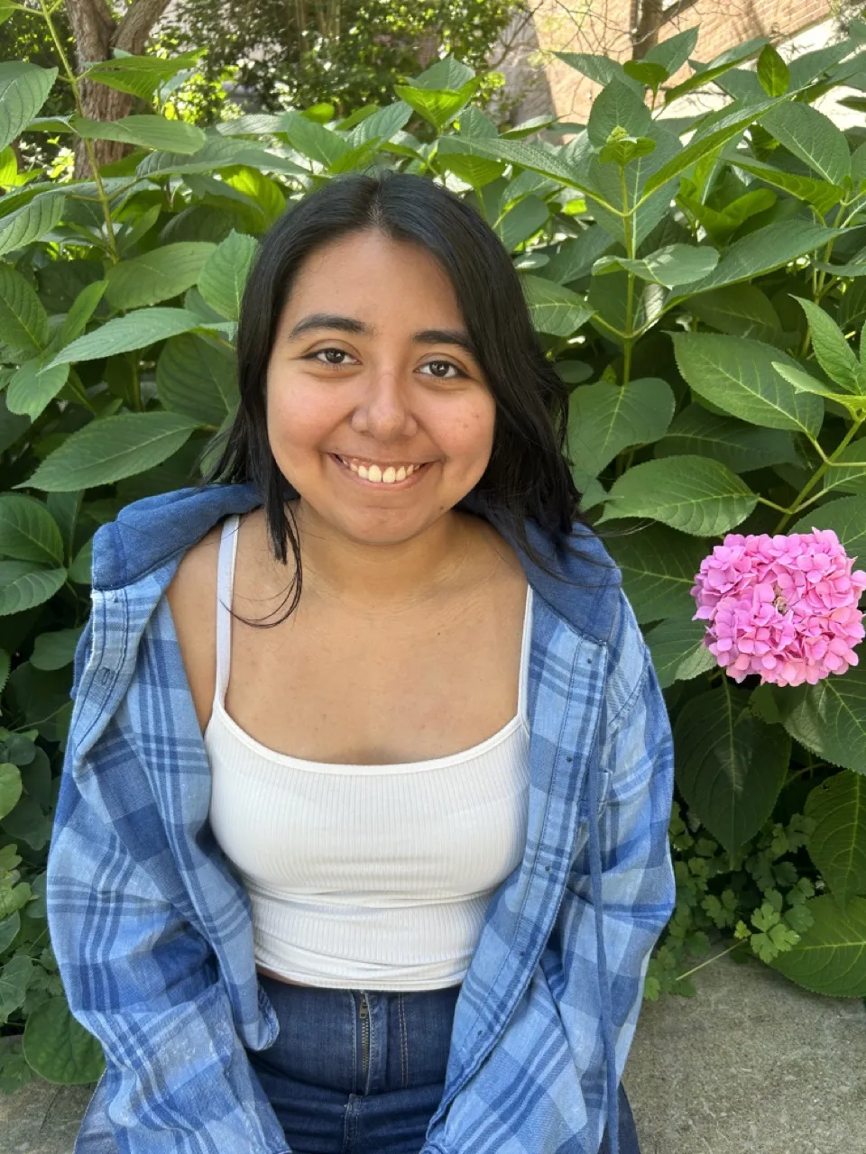 woman in front of bushes smiling, dark hair, blue plaid shirt