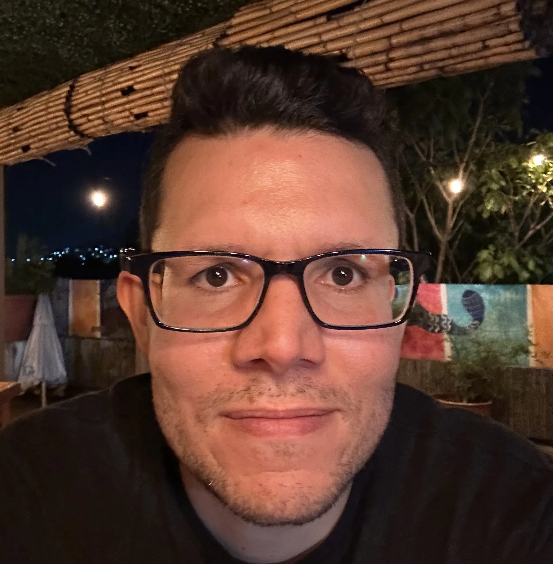 Image of a face with black glasses and a black shirt in a dark background.