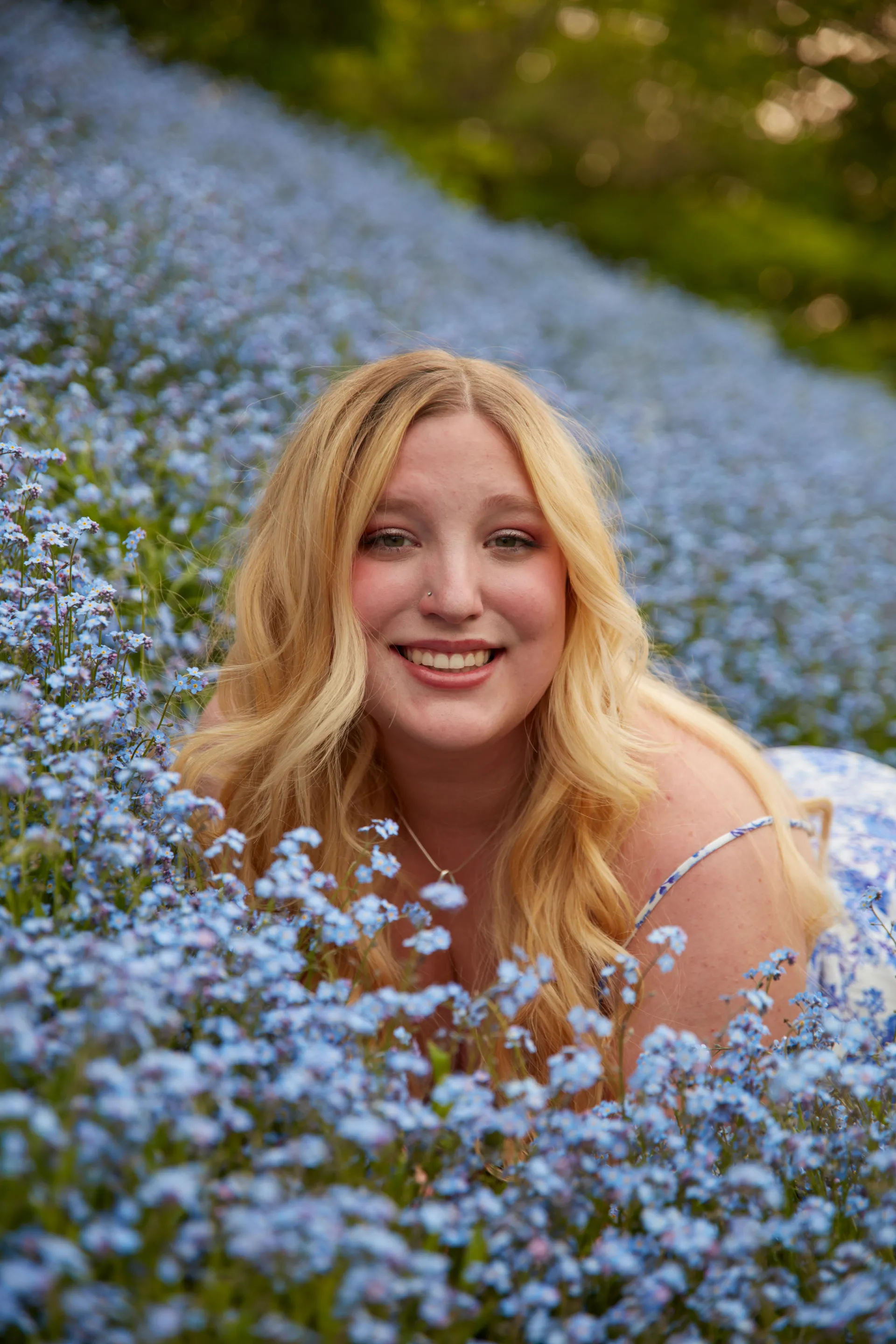 Younge, smiling, blonde woman lays in field of small blue flowers.