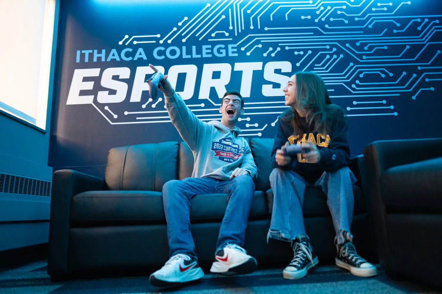 Two students play video games. The wall behind them says "Ithaca College Esports." 