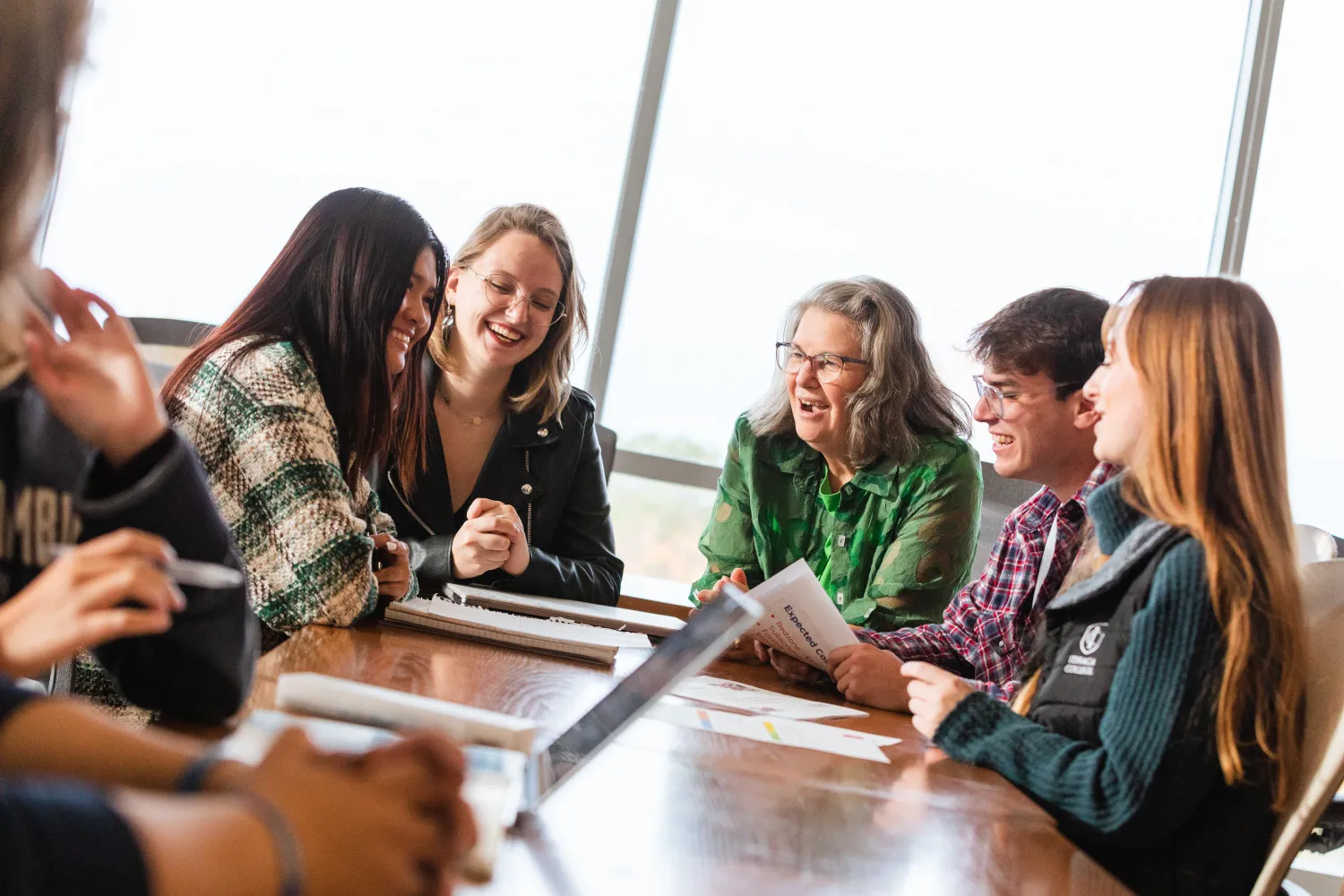 College students around a table laugh and smile with their professor. 