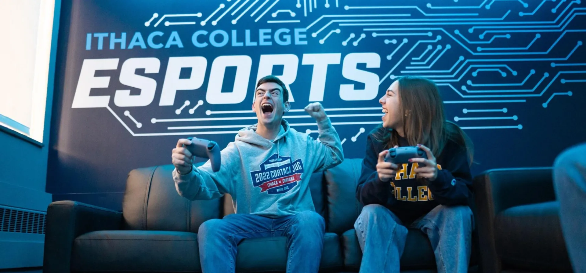 Two students sit on a couch holding PS5 controllers. They shout with excitement. On the wall behind them is the Ithaca College Esports Logo 