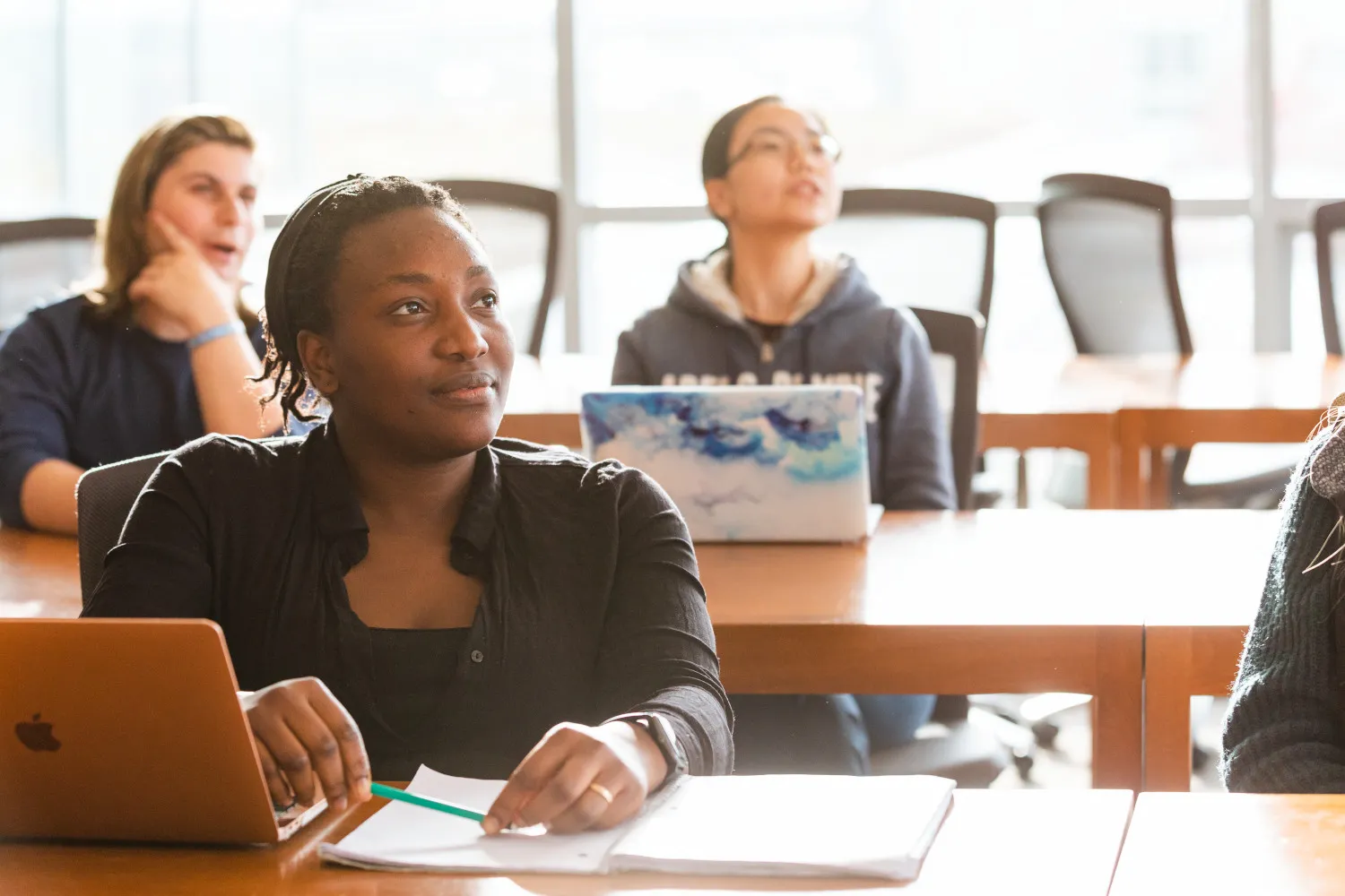 College students in a classroom; the student in the foreground has a thoughtful expression as she listens to a speaker out of frame. 