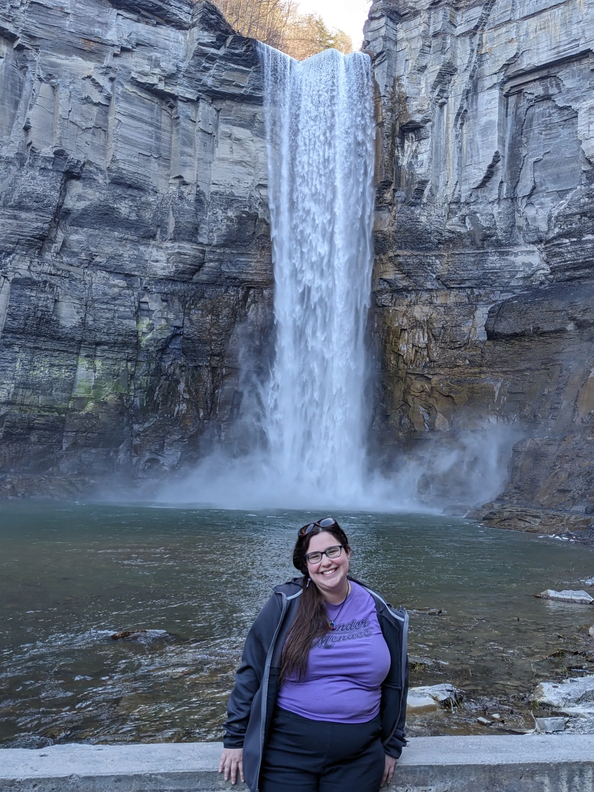 Images shows a smiling woman with long brown hair in a purple shirt in front of a tall and dramatic waterfall