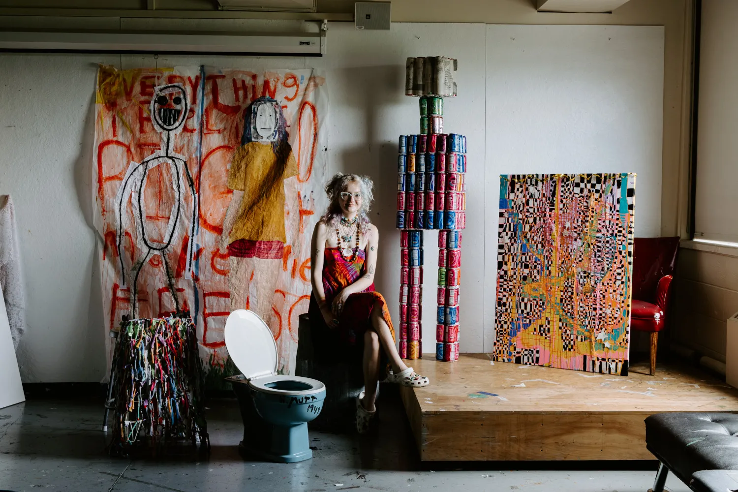 A student in an art studio is surrounded by the materials in their installation, including a giant robot made from discarded soda cans, a toilet, and original work. 
